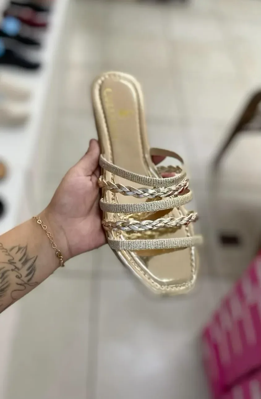 A rasteira dourada trançada segurada na mão, dentro da loja, com as caixas de sapato ao fundo